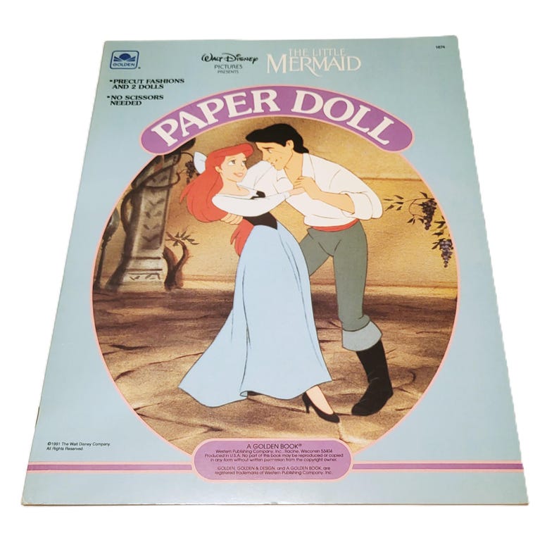 Pode incluir: Um livro de bonecas de papel vintage de "A Pequena Sereia". A capa apresenta Ariel e o Pr&iacute;ncipe Eric em um abra&ccedil;o rom&acirc;ntico. O livro &eacute; azul claro com o t&iacute;tulo "Paper Doll" em um oval rosa. O texto na capa diz "Precut Fashions and 2 Dolls" e "No Scissors Needed."