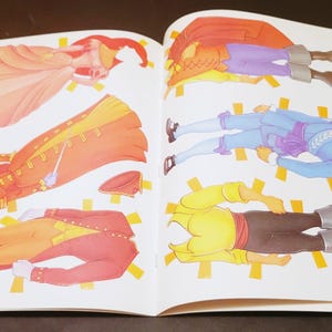 May include: Open book with colorful paper dolls. The dolls are dressed in historical costumes, including a long orange coat, a red coat, and a blue outfit. The paper dolls are cut out and ready to play.