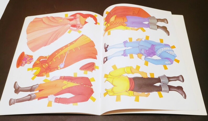 May include: Open book with colorful paper dolls. The dolls are dressed in historical costumes, including a long orange coat, a red coat, and a blue outfit. The paper dolls are cut out and ready to play.