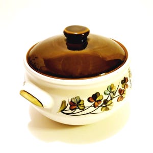May include: A brown ceramic casserole dish with a lid. The dish has a white background with a green, blue, and orange clover leaf design. The lid is brown and has a knob on top.