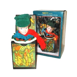 May include: A vintage musical jack-in-the-box featuring a boy wearing a green hat and red scarf. The box is decorated with a winter scene and the text "ENESCO presents TINY TIM action musical jack-in-the-box designed by artist Karen Habek".