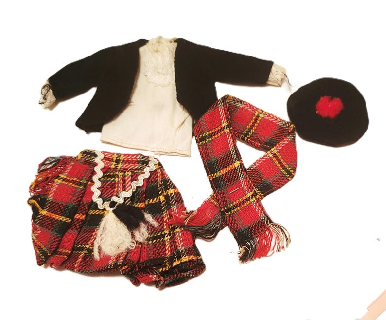 May include: A miniature Scottish outfit, featuring a black jacket, white shirt, red and black plaid kilt, scarf, and a black hat with a red pom-pom. The kilt has a white lace trim and a black and white tassel.
