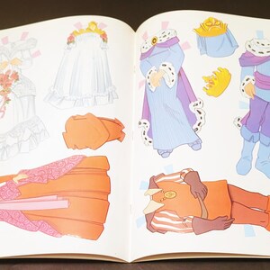 May include: An open book featuring paper dolls with various outfits. The dolls include a princess in a white gown, a queen in a purple robe, and a prince in a blue and purple outfit. Accessories include crowns and a bouquet.