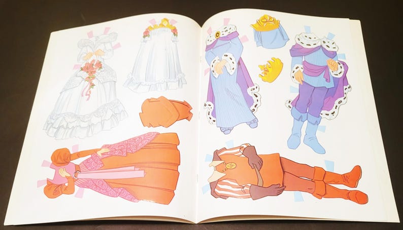 May include: An open book featuring paper dolls with various outfits. The dolls include a princess in a white gown, a queen in a purple robe, and a prince in a blue and purple outfit. Accessories include crowns and a bouquet.