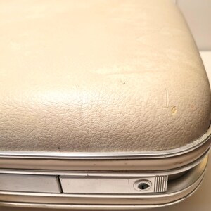 May include: Close-up of a vintage suitcase with a cream-colored, textured exterior. The suitcase has a silver-colored metal trim and a small lock. The surface shows signs of wear, including small scratches and imperfections.