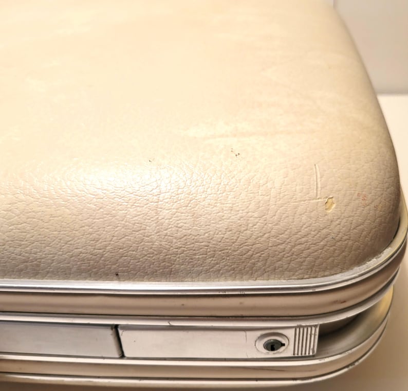 May include: Close-up of a vintage suitcase with a cream-colored, textured exterior. The suitcase has a silver-colored metal trim and a small lock. The surface shows signs of wear, including small scratches and imperfections.