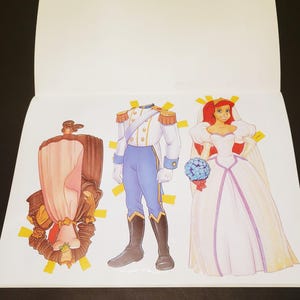 May include: A paper doll book featuring three illustrated figures. One figure is a brown and pink dress, another is a prince in a white and blue uniform, and the third is a bride in a white dress with red hair and a bouquet.