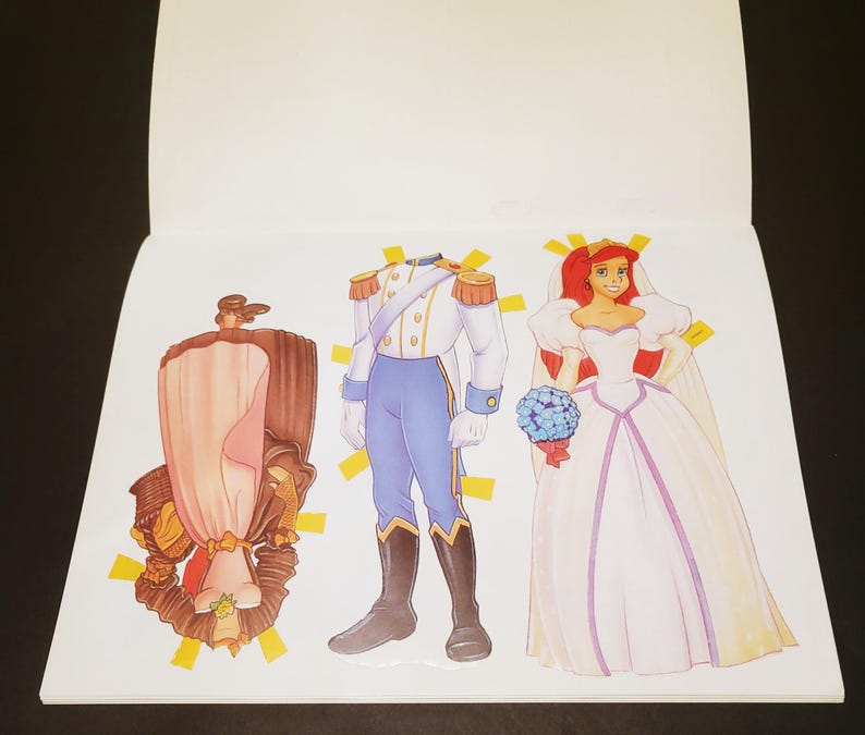 May include: A paper doll book featuring three illustrated figures. One figure is a brown and pink dress, another is a prince in a white and blue uniform, and the third is a bride in a white dress with red hair and a bouquet.