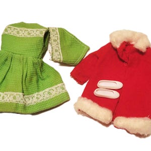 May include: A green doll dress and matching scarf with white lace trim, alongside a red coat with white faux fur trim and two white button closures. The doll clothing set is on a white background.