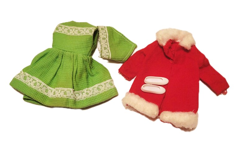 May include: A green doll dress and matching scarf with white lace trim, alongside a red coat with white faux fur trim and two white button closures. The doll clothing set is on a white background.