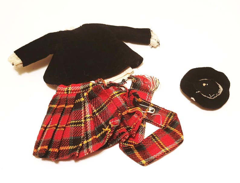 May include: A doll-sized school uniform set, featuring a black velvet jacket with lace trim, a red plaid pleated skirt, a matching tie, and a black hat with white embroidery. The set is designed for a doll.