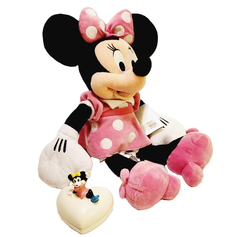 May include: A plush Minnie Mouse toy wearing a pink dress with white polka dots and a pink bow. The toy is sitting on a white heart-shaped box with a smaller Minnie Mouse toy inside.