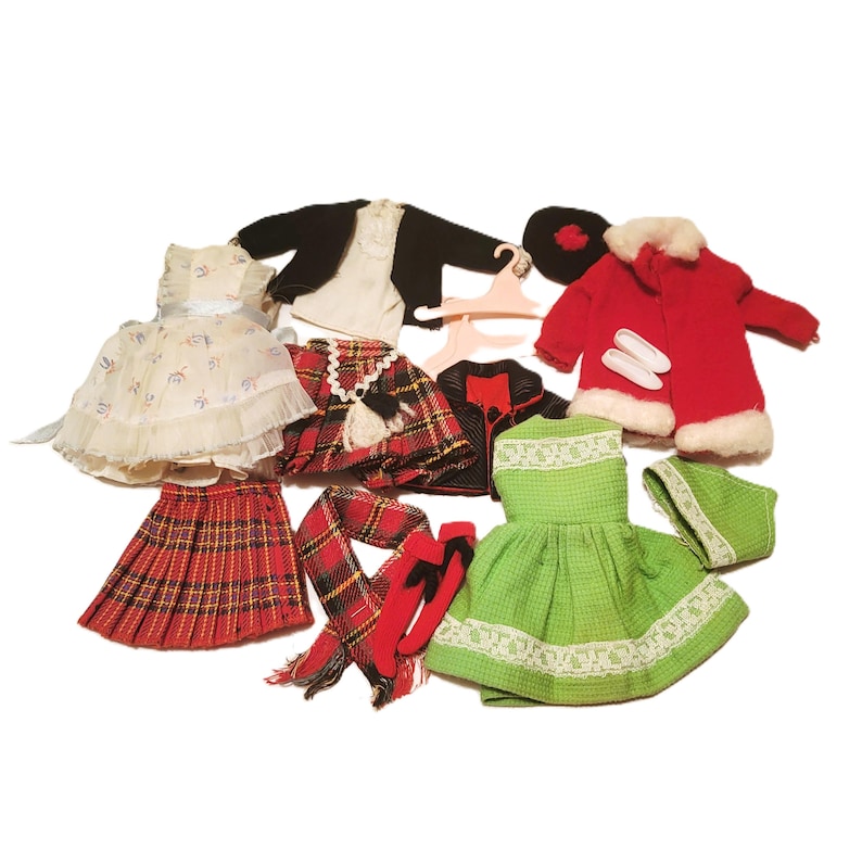 May include: A collection of vintage doll clothes, including a red plaid skirt, a green dress with lace trim, a red coat with white faux fur trim, and a black jacket. Other items include a scarf, hat, and shoes.