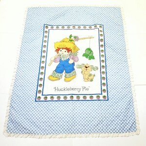 May include: A blue and white gingham baby blanket with a white border and a white ruffle. The center of the blanket features a colorful illustration of Huckleberry Pie, a cartoon character, fishing with a dog. The text "Huckleberry Pie" is printed below the illustration.