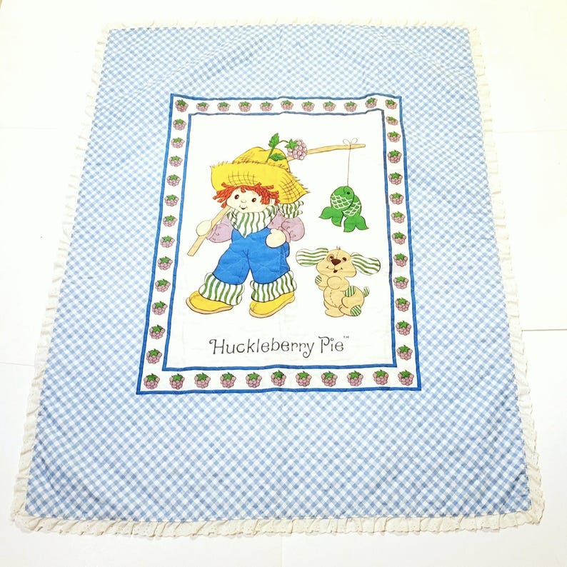 May include: A blue and white gingham baby blanket with a white border and a white ruffle. The center of the blanket features a colorful illustration of Huckleberry Pie, a cartoon character, fishing with a dog. The text "Huckleberry Pie" is printed below the illustration.