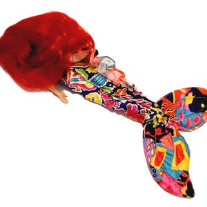 May include: A mermaid doll with long red hair and a colourful, patterned tail. The tail is made of fabric and has a variety of colours and designs.