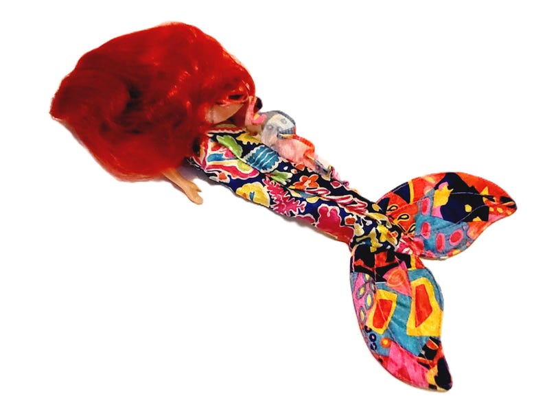 May include: A mermaid doll with long red hair and a colourful, patterned tail. The tail is made of fabric and has a variety of colours and designs.