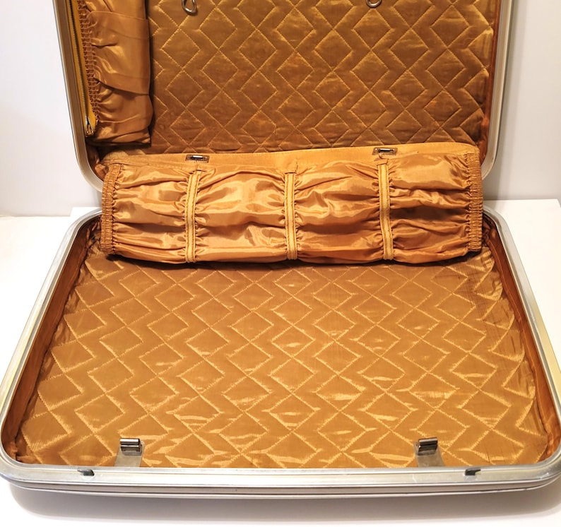May include: Open vintage suitcase with a silver frame and a gold quilted interior. The interior features a diamond pattern and several compartments, including a roll-up section. The suitcase is empty and ready for travel.