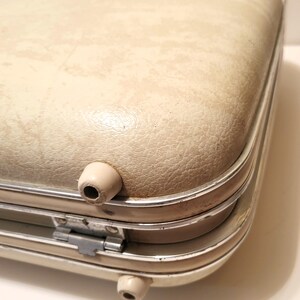 May include: Close-up of a vintage cream-colored suitcase with a textured surface. The suitcase has a silver trim and two beige feet. The metal hinge is visible. The suitcase is likely from the mid-century.