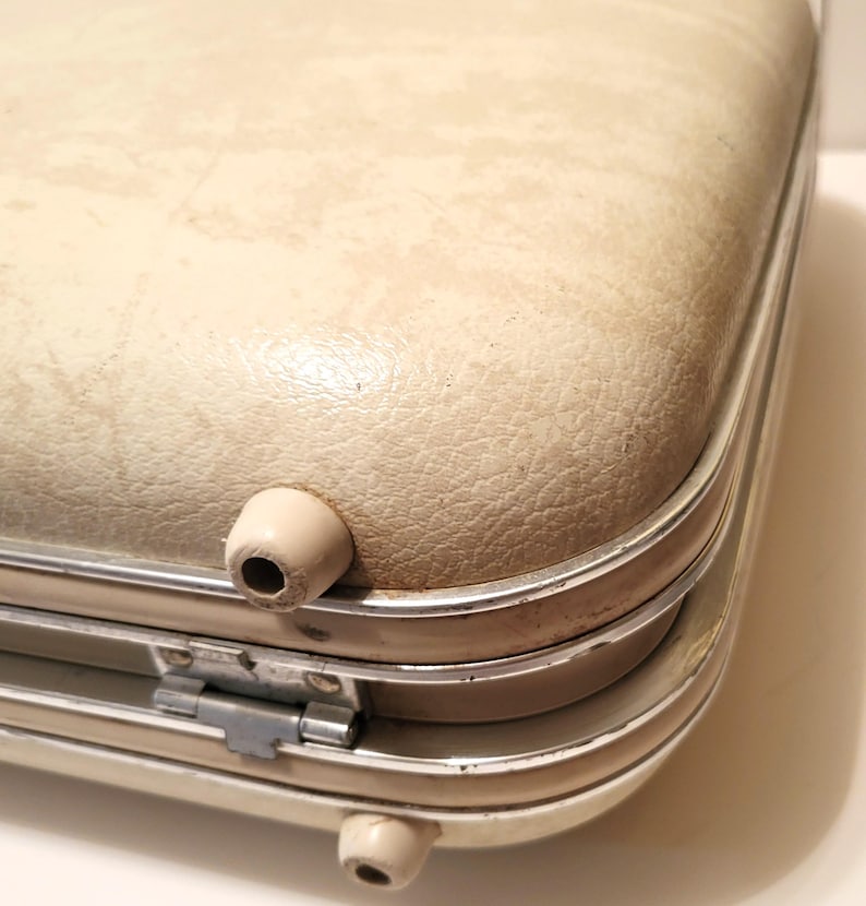 May include: Close-up of a vintage cream-colored suitcase with a textured surface. The suitcase has a silver trim and two beige feet. The metal hinge is visible. The suitcase is likely from the mid-century.