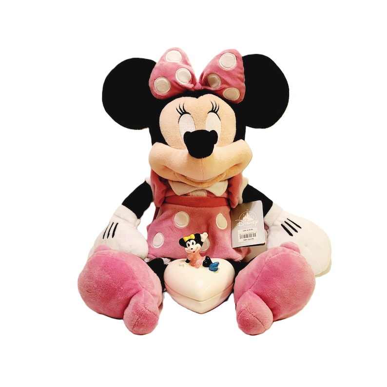 May include: A plush pink Minnie Mouse toy with a white polka dot dress and a red bow. She is holding a small white heart-shaped box with a miniature Minnie Mouse figurine inside.