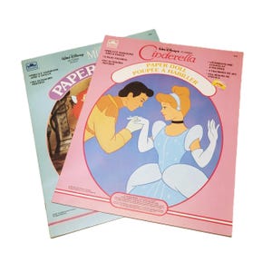 Pode incluir: Dois livros de bonecas de papel da Disney vintage. Um apresenta a Cinderela, com uma capa rosa e ilustra&ccedil;&otilde;es da Cinderela e do Pr&iacute;ncipe Encantado. O outro tem uma capa azul clara com uma imagem de uma princesa. Ambos os livros s&atilde;o rotulados como "Bonecas de papel".