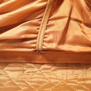 May include: Close-up of a golden-colored fabric with a quilted pattern at the bottom and a gathered, draped design above. The draped section has decorative trim with a rope-like texture. A small metal fastener is visible.