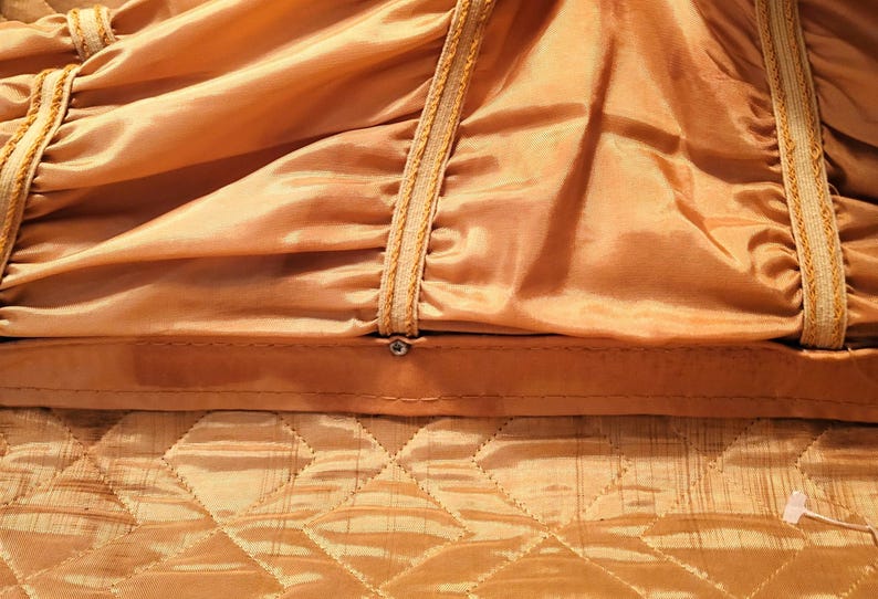 May include: Close-up of a golden-colored fabric with a quilted pattern at the bottom and a gathered, draped design above. The draped section has decorative trim with a rope-like texture. A small metal fastener is visible.
