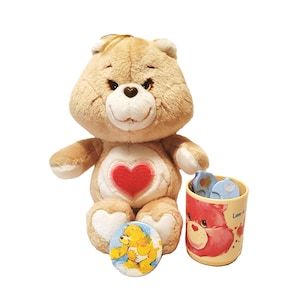 Peut inclure: Un ours en peluche beige avec un cœur rouge sur la poitrine, un badge rond jaune avec un ours de dessin animé et une petite tasse avec un ours rouge. La tasse contient de petits objets bleus et blancs. Sur fond blanc.