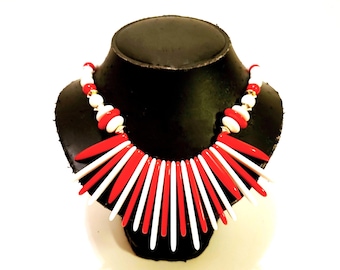 May include: A bold, statement necklace featuring alternating red and white pointed elements radiating from the center. The necklace has a black base and is displayed on a black mannequin bust. The necklace also has white and red beads.