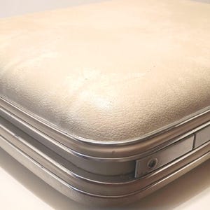 May include: Cream-colored vintage suitcase with a textured surface and silver-toned metal trim. The suitcase has a rounded top and a white handle. The case is closed and appears to be in good condition.
