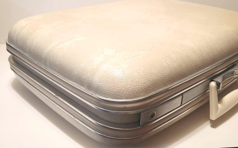 May include: Cream-colored vintage suitcase with a textured surface and silver-toned metal trim. The suitcase has a rounded top and a white handle. The case is closed and appears to be in good condition.