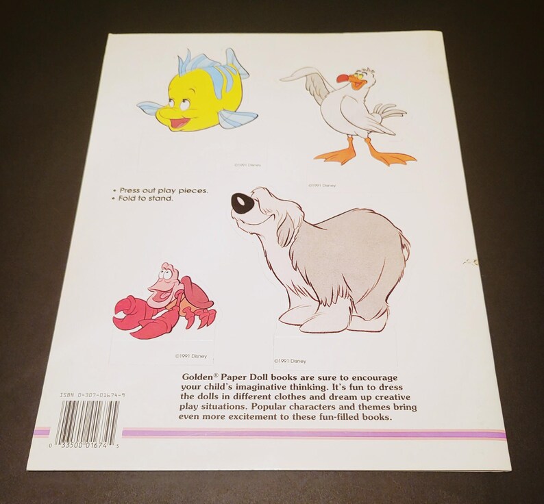 Pode incluir: Contra capa de um livro Golden Paper Doll com ilustra&ccedil;&otilde;es de personagens de desenhos animados. Inclui um peixe amarelo, uma gaivota branca, um caranguejo vermelho e um c&atilde;o cinzento. O texto diz "Press out play pieces. Fold to stand."