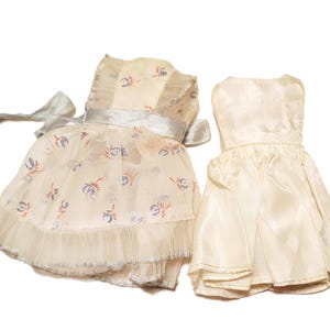 May include: Two doll dresses. One is a cream-colored dress with a floral pattern in blue and orange, a silver ribbon, and a ruffled hem. The other is a solid cream-colored dress with a pleated skirt.
