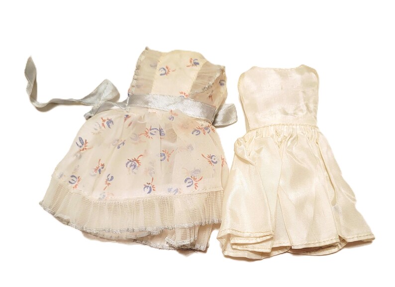 May include: Two doll dresses. One is a cream-colored dress with a floral pattern in blue and orange, a silver ribbon, and a ruffled hem. The other is a solid cream-colored dress with a pleated skirt.