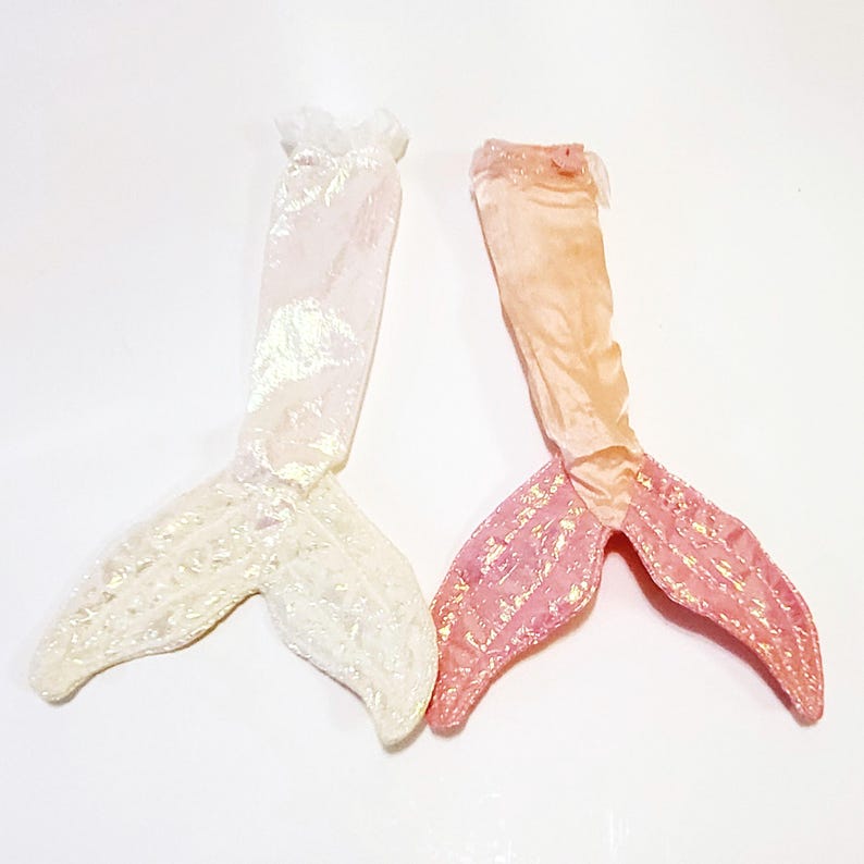 May include: Two mermaid tails made of fabric. One is white with iridescent shimmer and the other is pink with iridescent shimmer.