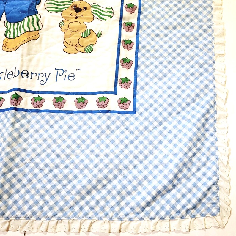 May include: A blue and white checkered baby blanket with a white ruffle border. The blanket features a cartoon illustration of a girl and a dog with the text "Huckleberry Pie" in a blue and white border.