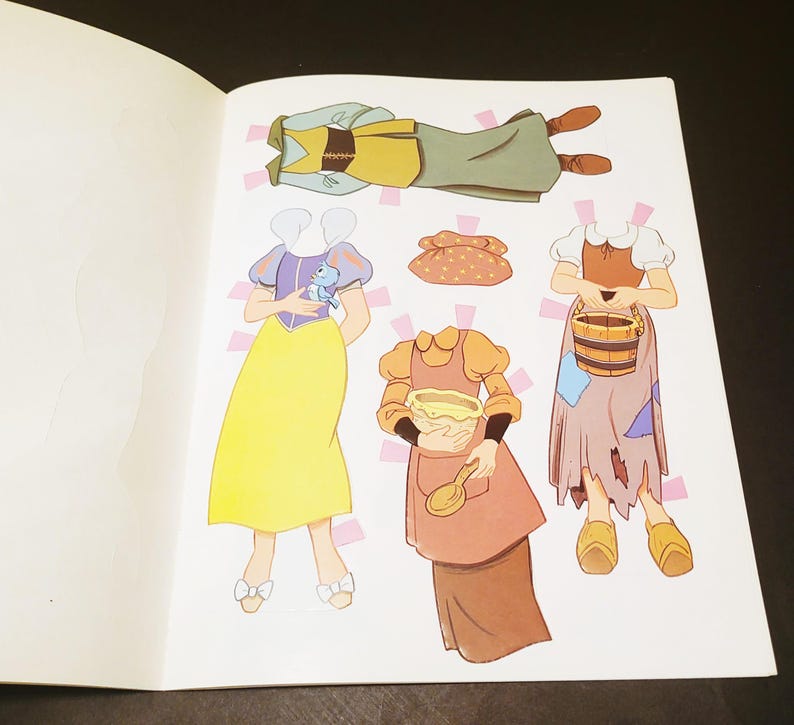 May include: A page from a children's paper doll book featuring illustrations of various outfits. The outfits include a yellow dress, a green and brown outfit, and a brown dress with an apron. The illustrations are colorful and whimsical.