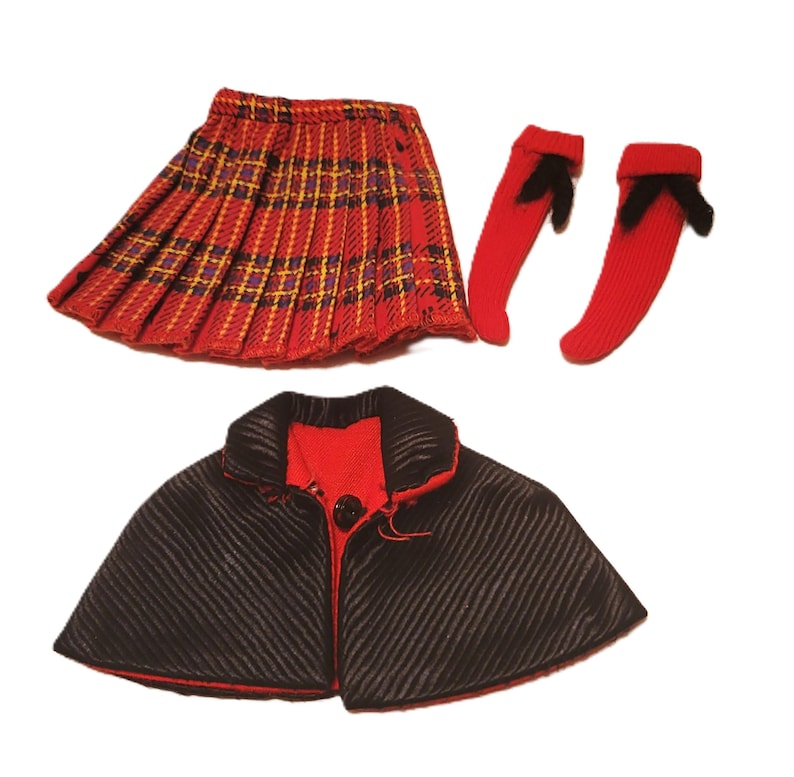 May include: A doll-sized outfit featuring a red plaid pleated skirt, red socks with black bows, and a black cape with red lining. The cape has a single button closure. The skirt has yellow and purple accents.