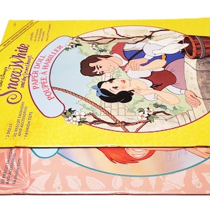 May include: Two vintage Golden Books paper doll sets. The top set features Snow White and the Seven Dwarfs, with a yellow background and illustrations of the characters. The bottom set has a pink background with an illustration of a mermaid.