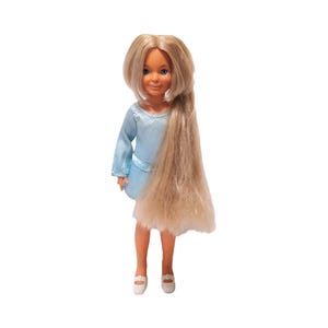 May include: A vintage doll with long blonde hair and blue eyes. The doll is wearing a light blue dress and white shoes. The doll's hair is styled with a side part and a long, flowing ponytail. The doll is standing on a white background.