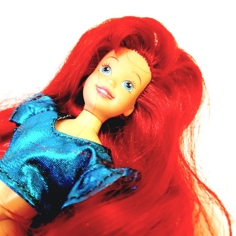 May include: A doll with red hair and blue eyes, wearing a blue sparkly top. The doll is looking upwards and smiling.