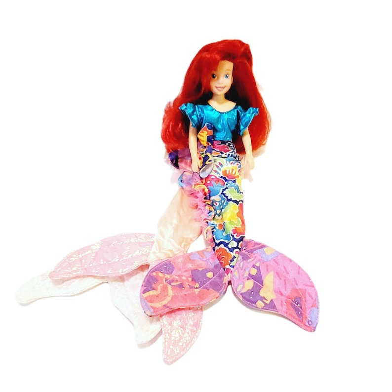 May include: A red-haired mermaid doll with a blue top and a colourful, patterned tail. The doll is sitting on a white background.