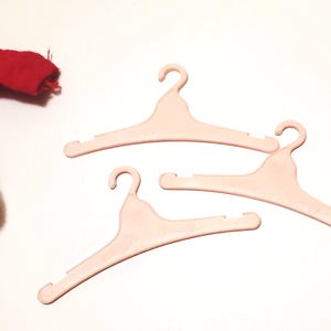 May include: Three light pink plastic clothes hangers with curved hooks and notched shoulders. A red garment with white trim is partially visible in the upper left corner. The hangers are on a white surface.