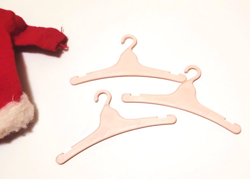 May include: Three light pink plastic clothes hangers with curved hooks and notched shoulders. A red garment with white trim is partially visible in the upper left corner. The hangers are on a white surface.
