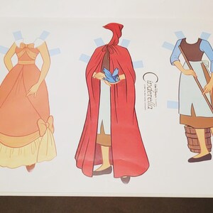 Pode incluir: Livro de bonecas de papel com tr&ecirc;s figuras ilustradas: uma princesa com um vestido cor de p&ecirc;ssego, uma figura com uma capa vermelha com capuz e Cinderela com uma roupa azul e castanha. O texto "Cinderella" &eacute; vis&iacute;vel na p&aacute;gina.