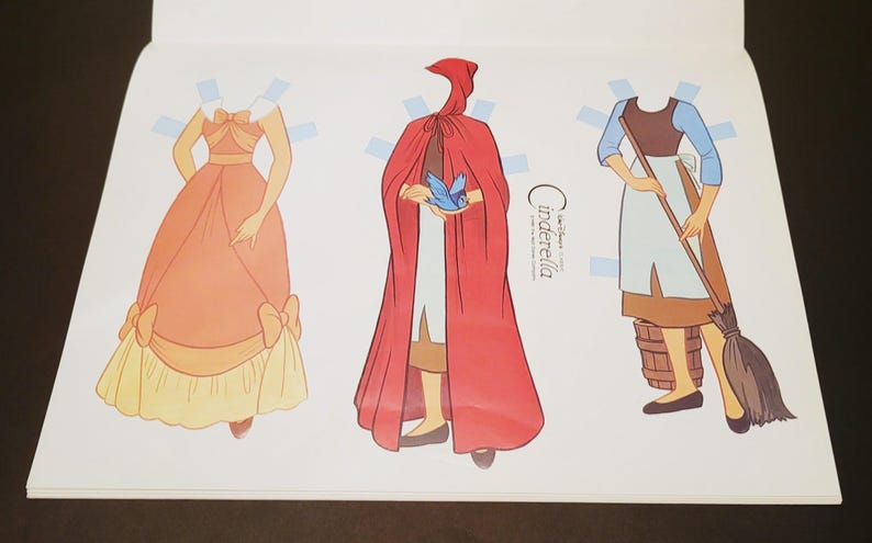 Pode incluir: Livro de bonecas de papel com tr&ecirc;s figuras ilustradas: uma princesa com um vestido cor de p&ecirc;ssego, uma figura com uma capa vermelha com capuz e Cinderela com uma roupa azul e castanha. O texto "Cinderella" &eacute; vis&iacute;vel na p&aacute;gina.