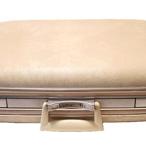 May include: A vintage beige suitcase with a rounded top and silver trim. The suitcase has a cream-colored handle and a metal clasp with the word "Jetters" on it. The suitcase is closed and appears to be in good condition.