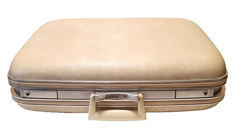 May include: A vintage beige suitcase with a rounded top and silver trim. The suitcase has a cream-colored handle and a metal clasp with the word "Jetters" on it. The suitcase is closed and appears to be in good condition.