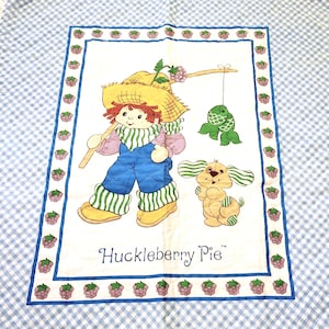 May include: A white and blue gingham patterned blanket with a framed image of Huckleberry Pie, a red-haired girl in a straw hat, holding a fishing pole with a green fish on the line. A yellow dog is standing next to her. The frame is decorated with a border of purple raspberries.